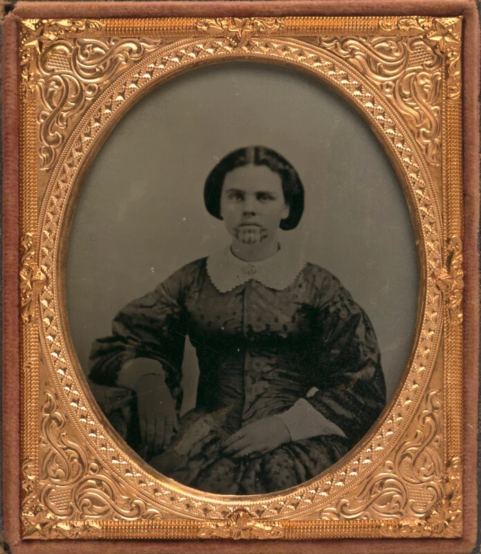 A daguerreotype portrait of a woman with a chin tattoo, wearing a dark patterned dress with a white collar, set in an ornate gold oval frame.