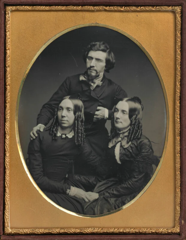 A daguerreotype portrait of a bearded man standing behind two seated women, all dressed in mid-19th-century formal attire.