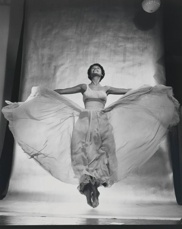 A woman with short dark hair jumps in the air, wearing a flowing, light-colored gown that spreads out around her against a plain backdrop.