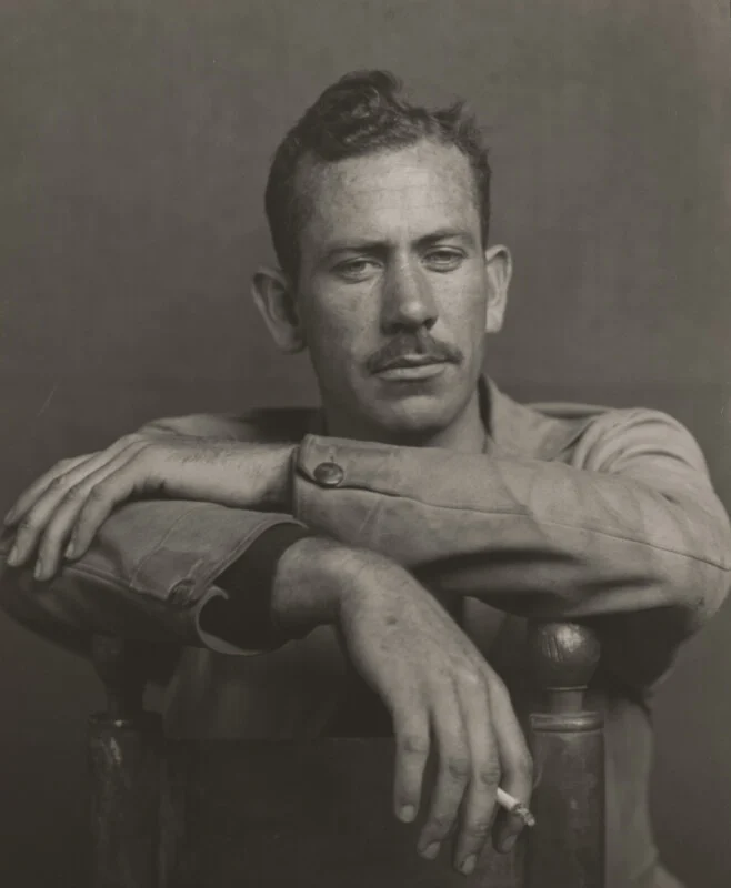 A black-and-white portrait of a man with a mustache, leaning his arms on the back of a chair while holding a cigarette.