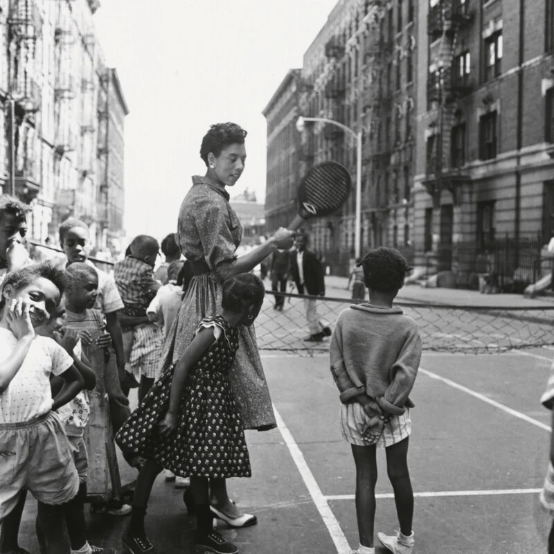 A woman holding a tennis racket stands on a city street court surrounded by a group of children, with apartment buildings in the background.