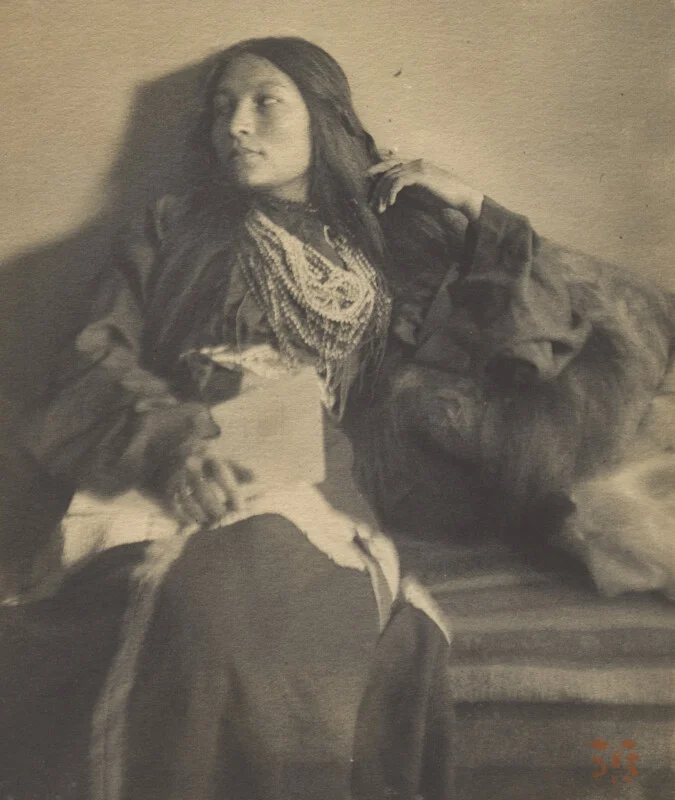 A portrait of a young woman with long dark hair, wearing a layered beaded necklace and a dark garment, seated against a wall.