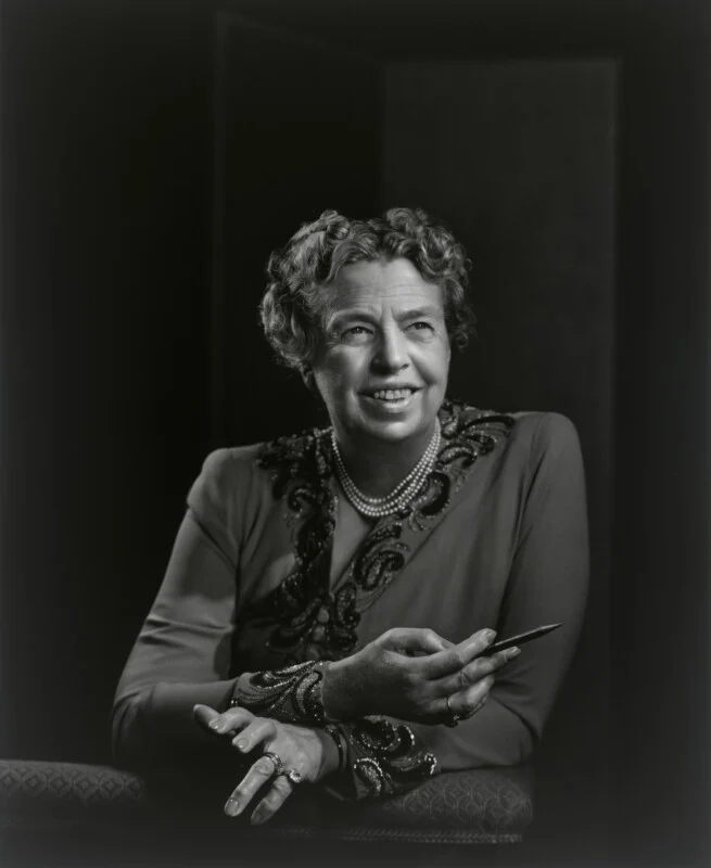 A black-and-white portrait of Eleanor Roosevelt, wearing a beaded dress and pearls, holding a pencil while looking to the side.