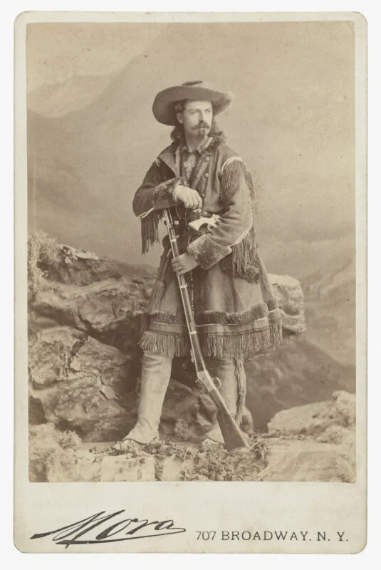 A studio portrait of William "Buffalo Bill" Cody standing on a rock formation, wearing a fringed buckskin coat and holding a rifle.