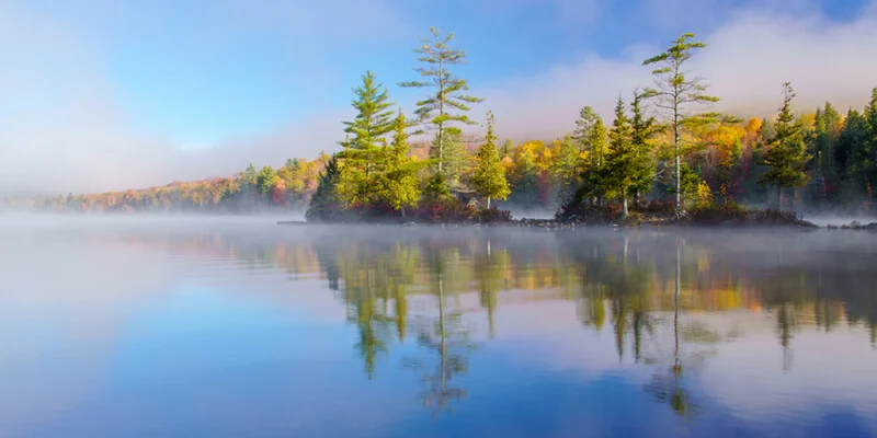 A calm lake reflects a small, tree-covered island surrounded by morning mist and autumn-colored foliage.