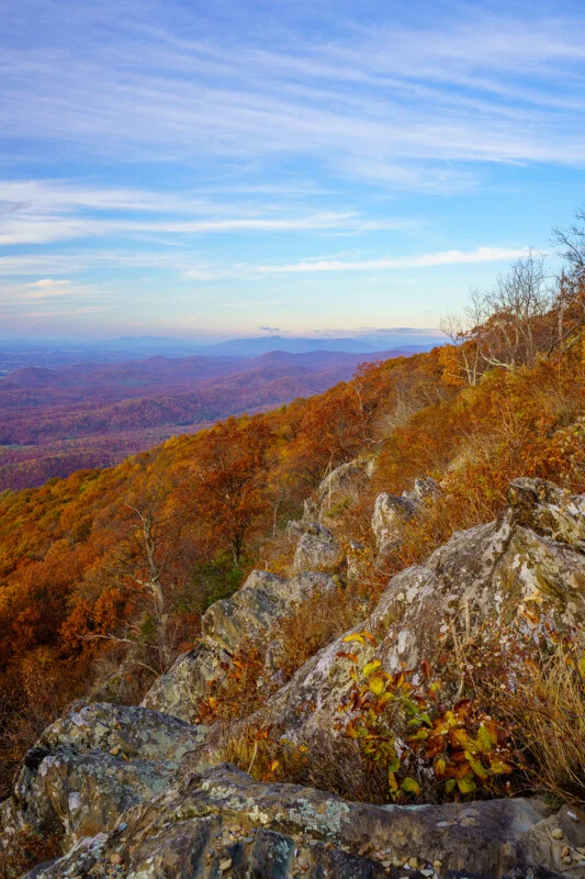 A rocky, autumn-colored mountainside overlooks a vast, hazy valley under a blue sky with wispy clouds.