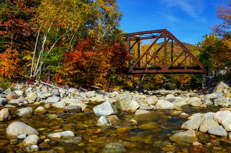 A rusty steel truss bridge spans a shallow, rocky stream surrounded by trees with vibrant autumn foliage under a clear blue sky.