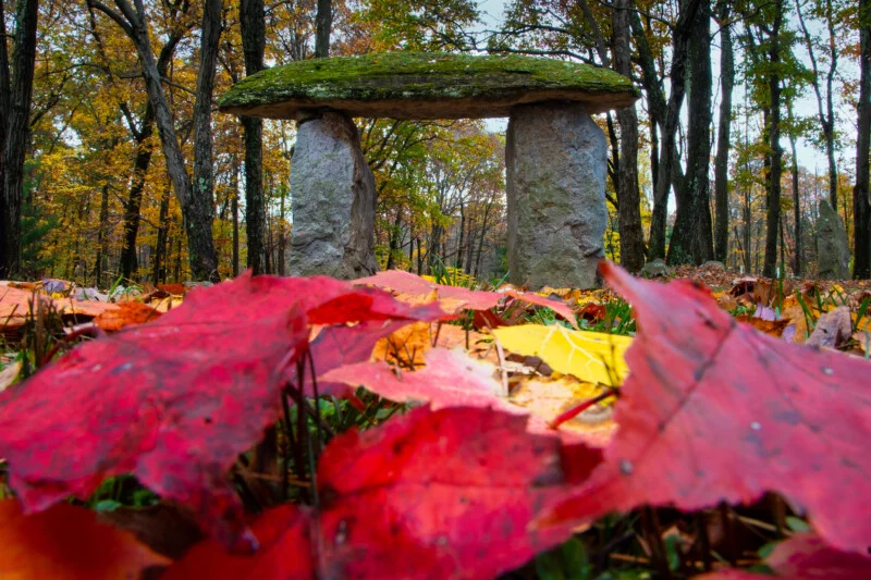 A moss-covered stone slab rests atop two upright stone pillars in a forest filled with colorful autumn leaves.