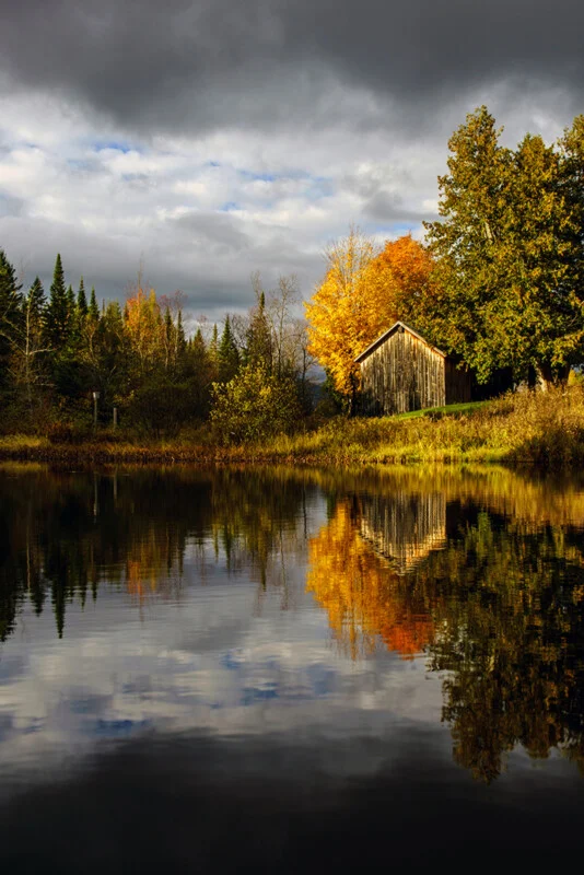 A rustic wooden barn sits beside a calm pond, surrounded by autumn trees under a cloudy, dramatic sky.