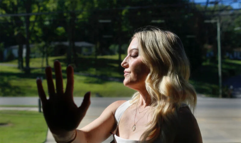 A blonde woman in a white top stands in profile with her eyes closed, holding her hand up toward the light.
