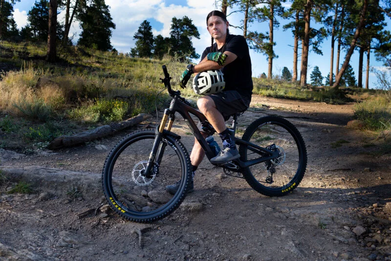 A man with long braided hair sits on a mountain bike on a dirt trail in a forest, holding a helmet under his arm.