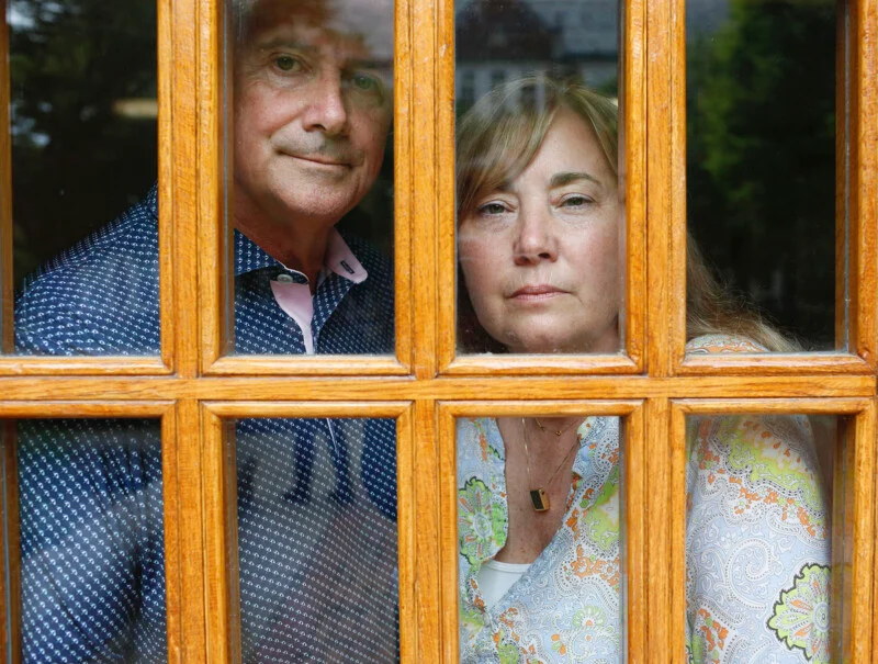 A man and a woman stand side-by-side behind a wooden-framed glass door, looking directly at the camera.