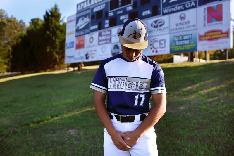A young baseball player in a blue and white Wildcats uniform with the number 17 stands with his head bowed in front of a scoreboard.