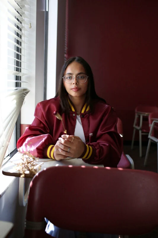 A young person with glasses and a maroon varsity jacket sits at a desk by a window, holding a dried flower.