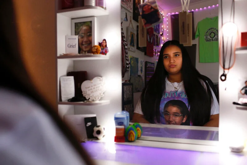 A young woman with long dark hair sits at a vanity mirror, wearing a shirt featuring a portrait of a girl in glasses.