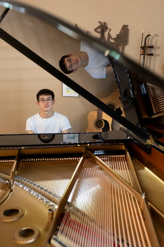 A young man with glasses and a white t-shirt sits at a grand piano, his reflection visible in the instrument's polished lid.