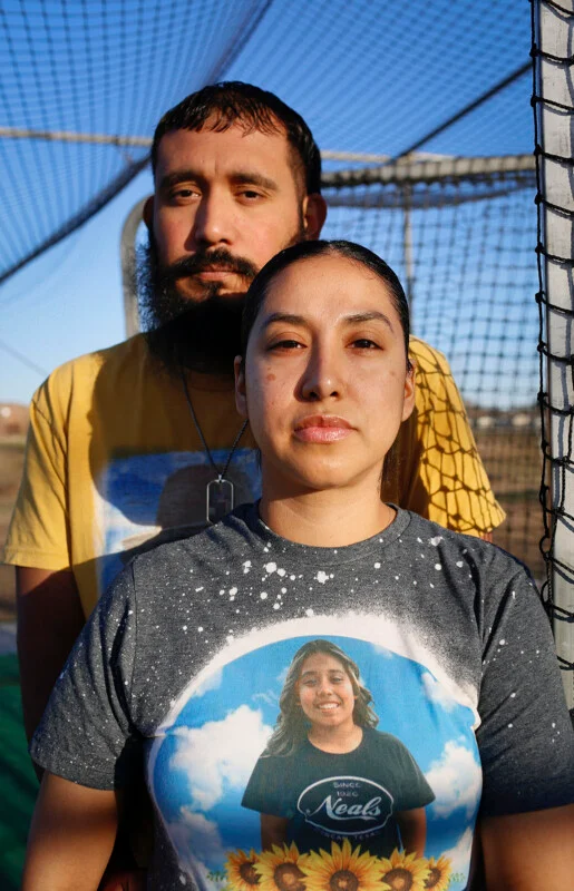 A man with a beard and a woman stand close together in front of a sports net, with the woman wearing a shirt featuring a portrait of a smiling girl.