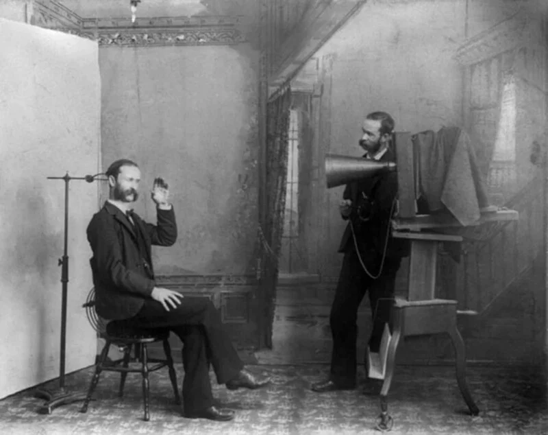 A man sits in a chair with his head held steady by a metal stand while a photographer operates a large, bellows-style camera on a tripod.