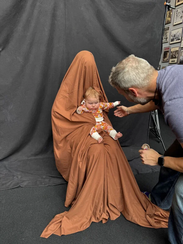A man in a gray t-shirt interacts with a baby sitting on the lap of a person completely covered by a brown fabric drape.