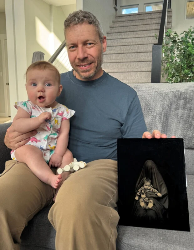 A man sits on a couch holding a baby on his lap while displaying a framed black-and-white portrait of the same baby.
