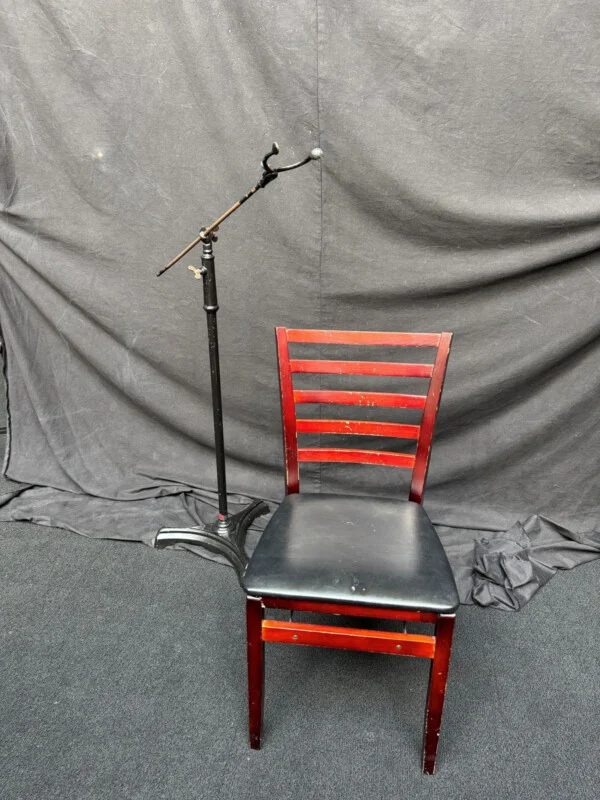 A black metal stand with an adjustable arm sits next to a wooden chair with a black padded seat and a ladder-back design against a dark fabric backdrop.
