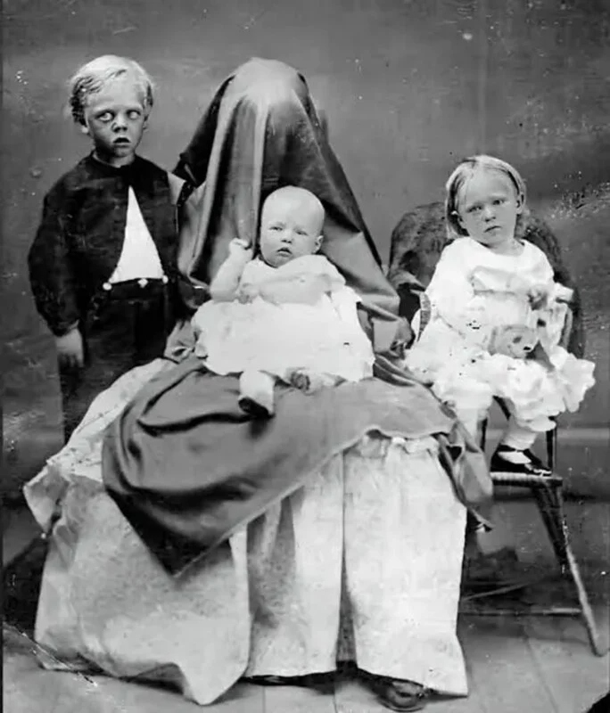 A vintage black-and-white portrait of a mother covered by a dark cloth, holding a baby, flanked by a young boy and girl.