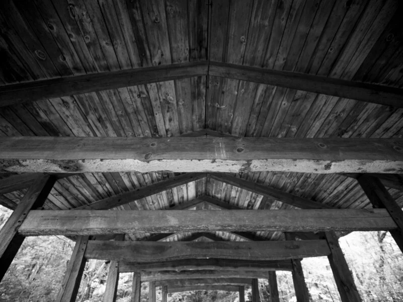 A low-angle, black-and-white view looking up at the wooden rafters and plank ceiling of an open-sided structure.