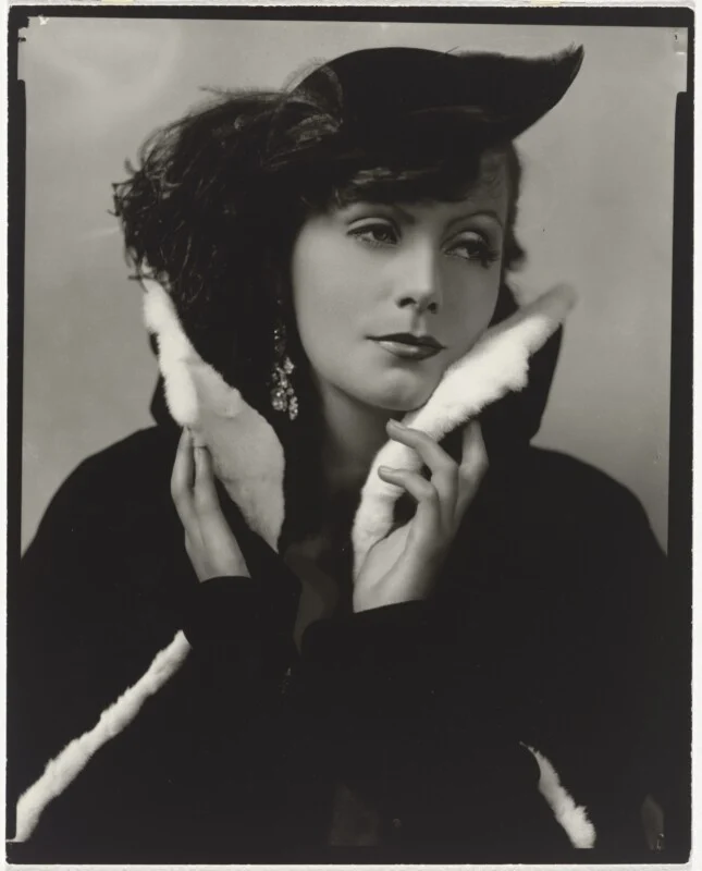 A black-and-white portrait of Greta Garbo wearing a dark coat with a white fur collar and a feathered hat, holding the collar up to her chin.