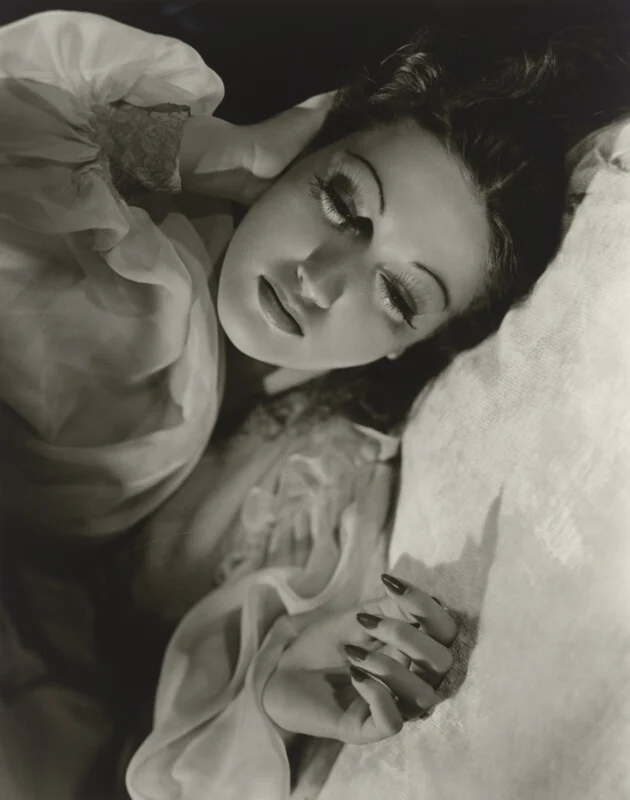 A black-and-white portrait of a woman with long eyelashes lying down, resting her head on her hand.