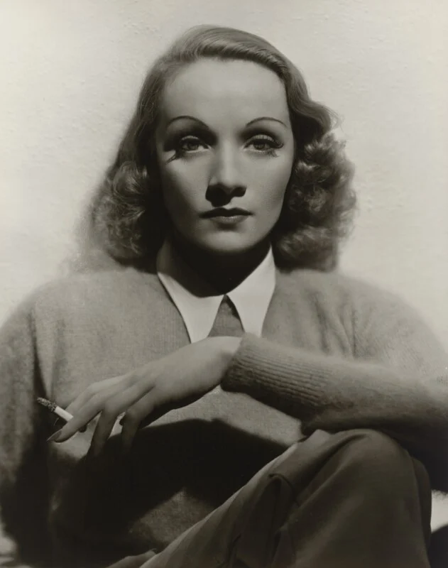 A black-and-white portrait of Marlene Dietrich wearing a collared shirt and sweater, holding a cigarette in her right hand.