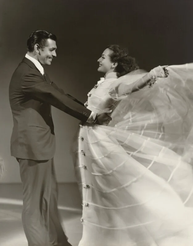 Clark Gable and Joan Crawford dance together, with Crawford wearing a flowing white gown that billows out to the side.