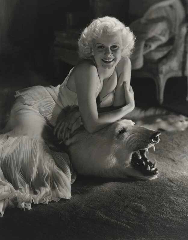 A blonde woman in a light-colored, pleated gown smiles while resting her arms on the head of a taxidermied polar bear rug.