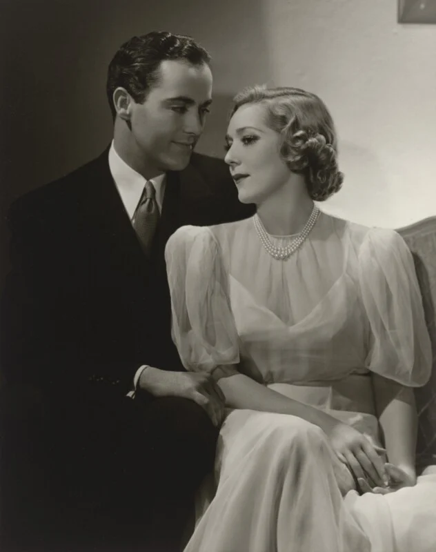 A man in a dark suit and tie sits beside a woman in a light-colored, short-sleeved gown and pearl necklace.