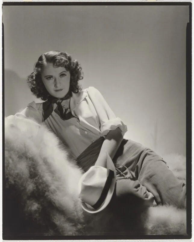 A woman with wavy brown hair, wearing a white collared shirt, a dark neckerchief, and light-colored trousers, reclines on a fur-covered surface while holding a white fedora.