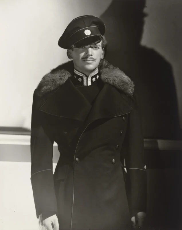 A man with a thin mustache wears a dark, fur-collared military-style coat and a peaked cap with a circular emblem.