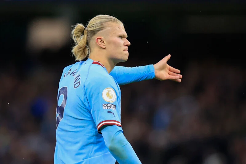 Erling Haaland, wearing a light blue Manchester City jersey with the number 9, gestures with his right hand while looking to the side.