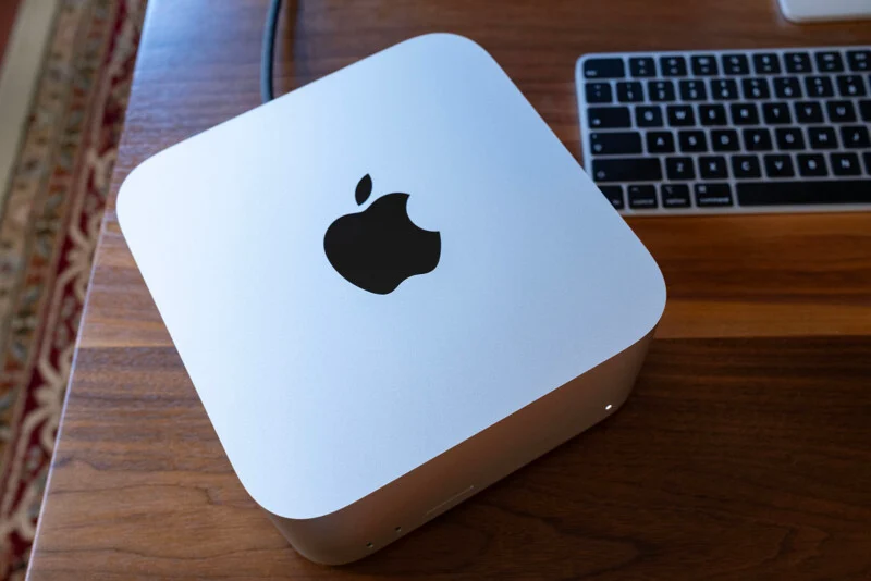 A silver Apple Mac mini sits on a wooden desk next to a wireless keyboard.