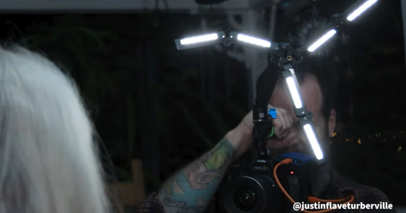 A person with a tattooed arm holds a camera equipped with a multi-armed LED light rig, filming a person with long white hair.