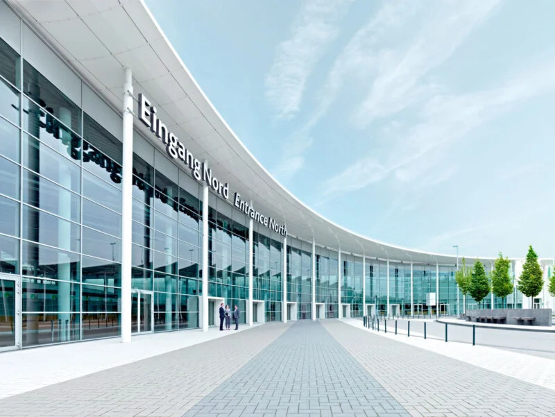 A modern, curved glass building with the sign "Eingang Nord Entrance North" features a wide paved walkway leading to its entrance, where three people stand.