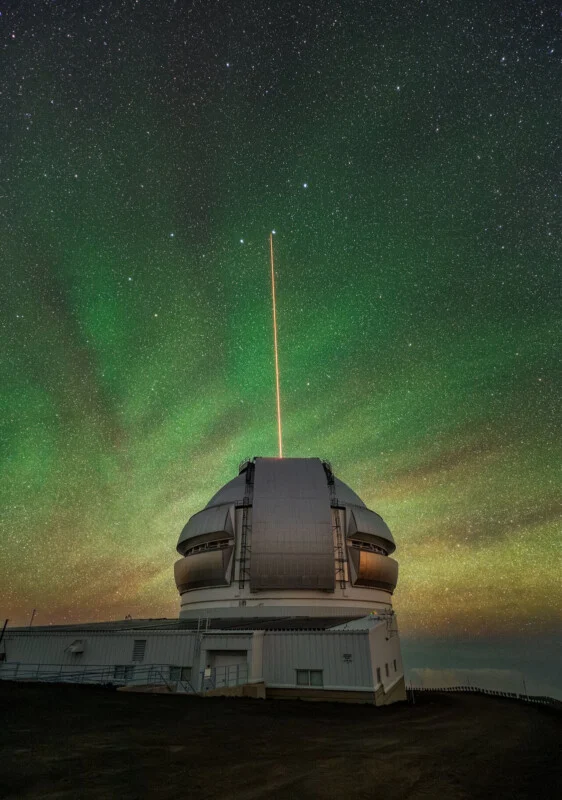 A large observatory dome sits under a starry night sky with a green airglow, emitting a bright orange laser beam upward.